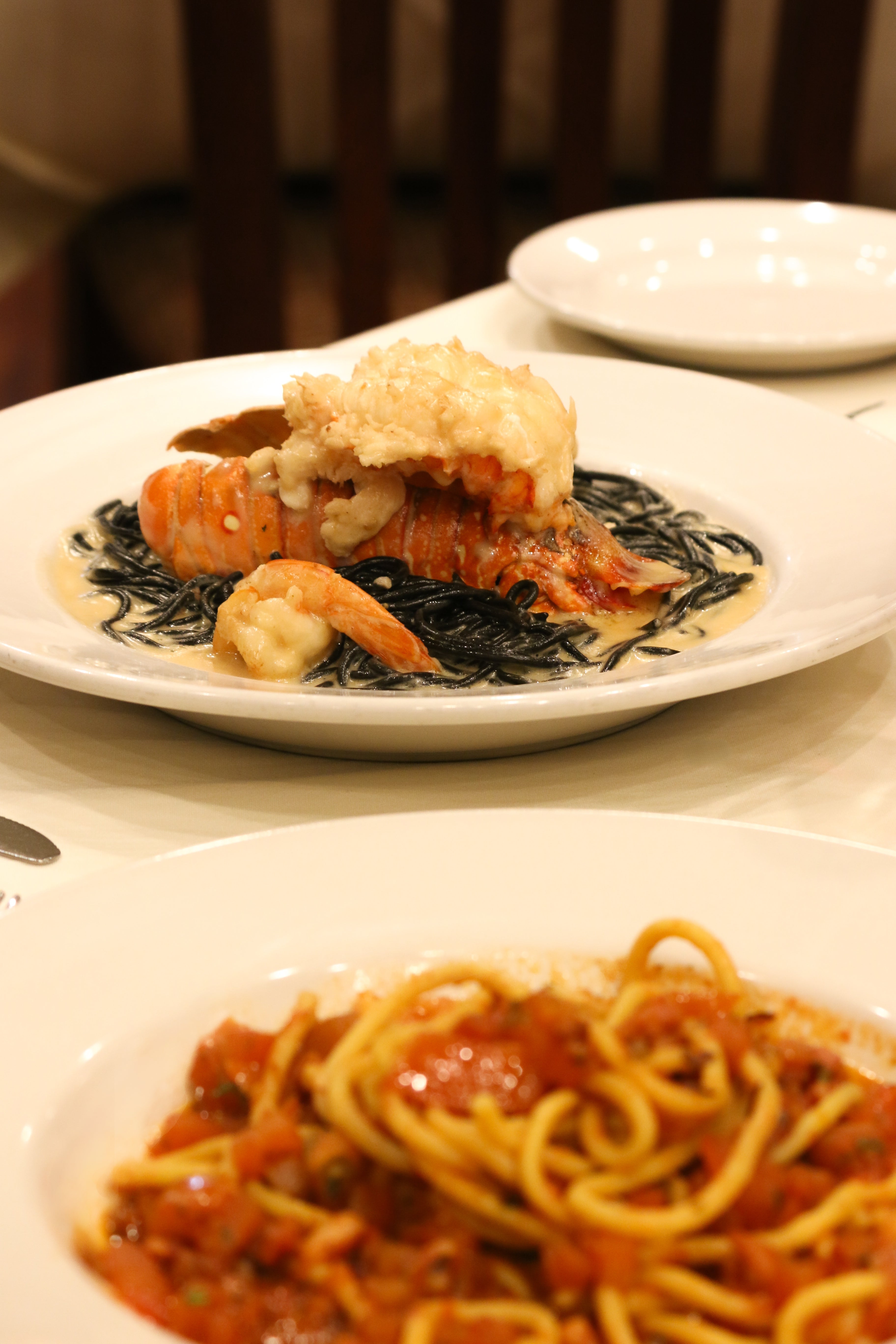 A photo of two plates of pasta from Anacapri Italian restaurant. The first plate features lobster and squid ink noodles, while the second plate contains bucatini pasta with red sauce.